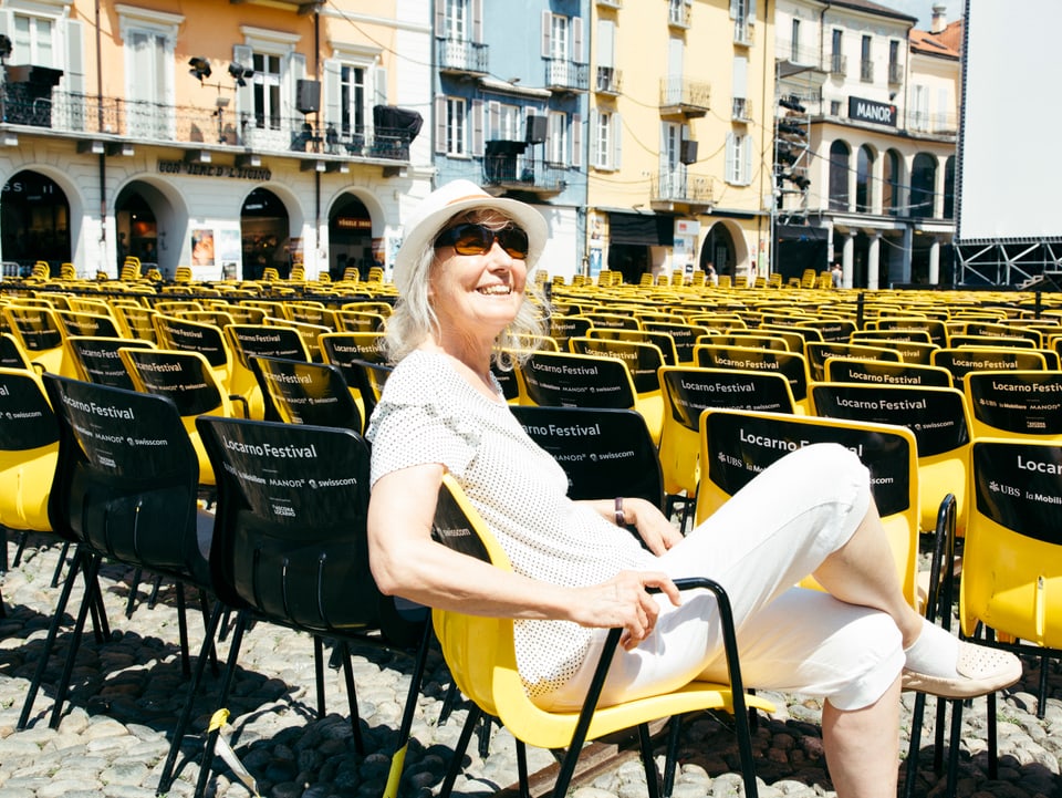 Ältere Frau mit Hut und Sonnenbrille sitzt lachend in einem gelben Plastikstuhl