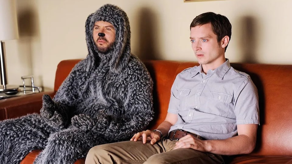 Mann im Hundekostüm sitzt neben einem Mann auf dem Sofa