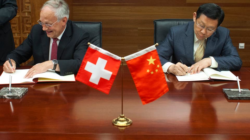 Johann Schneider-Ammann und der chinesische Handelsminister Chen Deming unterzeichnen Verträge. Im Vordergrund Schweizer und chinesische Fähnchen.