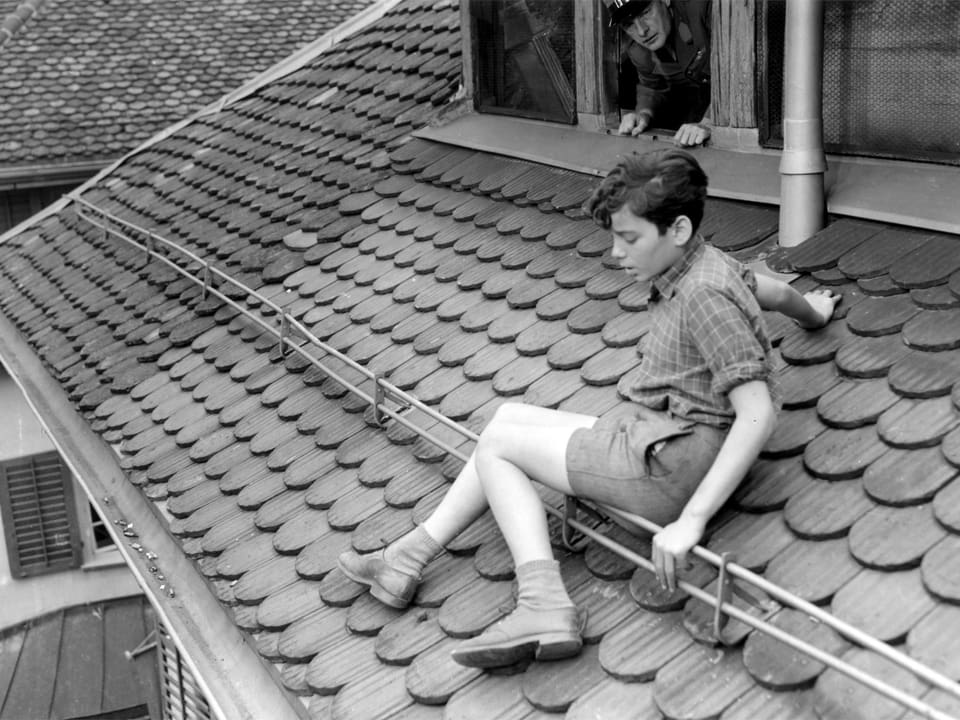 Ein Jugendlicher sitzt auf einem Ziegeldach und blickt in die Tiefe. Hinter ihm scheint ein Wachtmeister aus einem Dachfenster auf ihn einzureden.