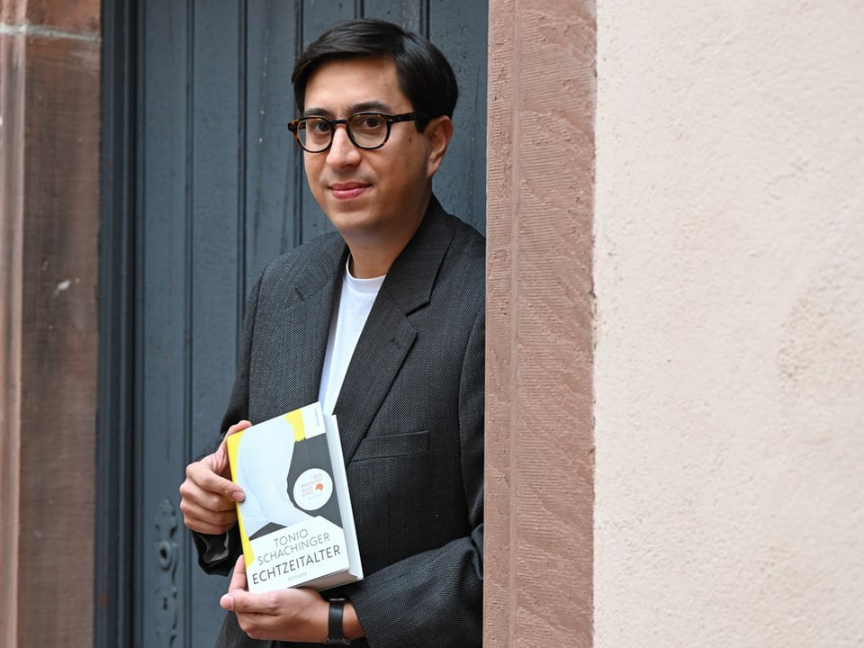 Ein Mann mit dunkler Brille und Anzug steht vor einer Tür und hält ein Buch hoch. 