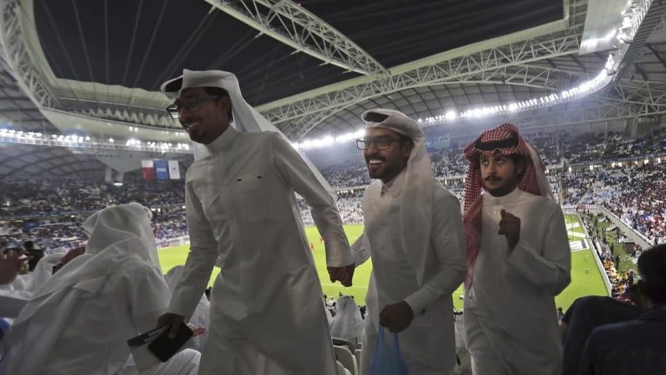 Drei Männer laufen nebeneinander im Gang auf einer Stadiontribüne. Im Hintergrund läuft ein Fussballspiel.