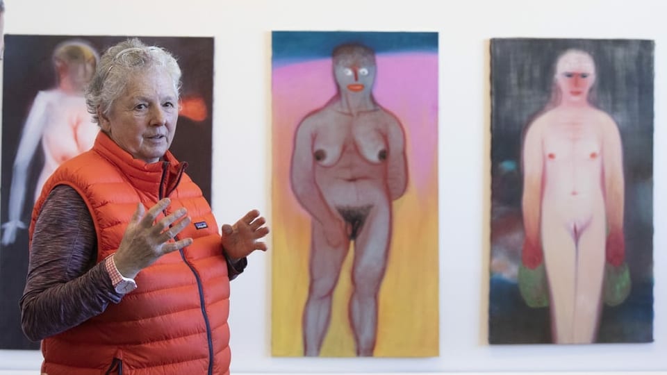 Frau mit grauen Haaren und oranger Weste steht im Museum vor lebensgrossen Bildern von nackten Frauen