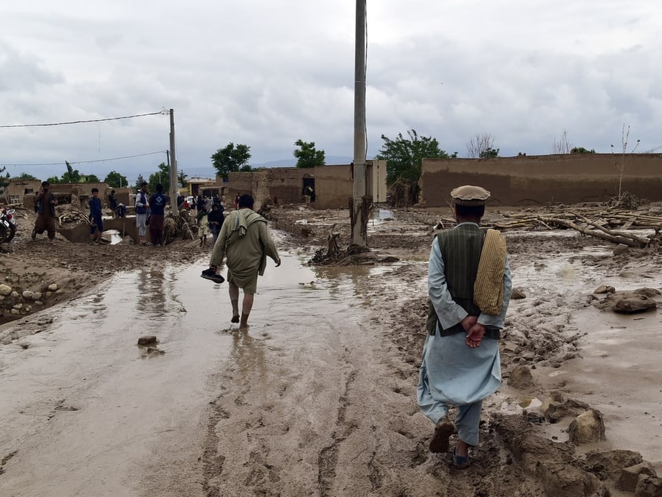 Menschen gehen durch ein überflutetes Dorf mit zerstörten Lehmgebäuden.