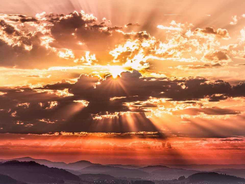 Tief stehende Sonne bei gelbem bis rotem Himmel, davor einige Wolken und Sonne wirft Strahlen. 