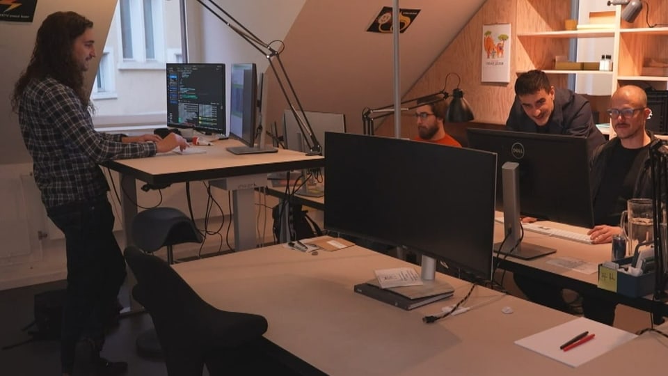 Vier Personen arbeiten in einem Büro. Zwei Männer besprechen etwas zusammen und blicken in einen Desktop.