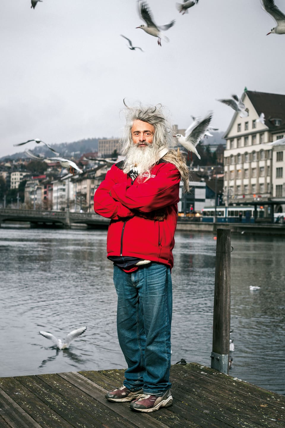Ein Matt mit langem, zotteligen, weissen Haar, einer roten Windjacke, blauen Jeans und Turnschuhen. Er steht auf einem Steg in Zürich.