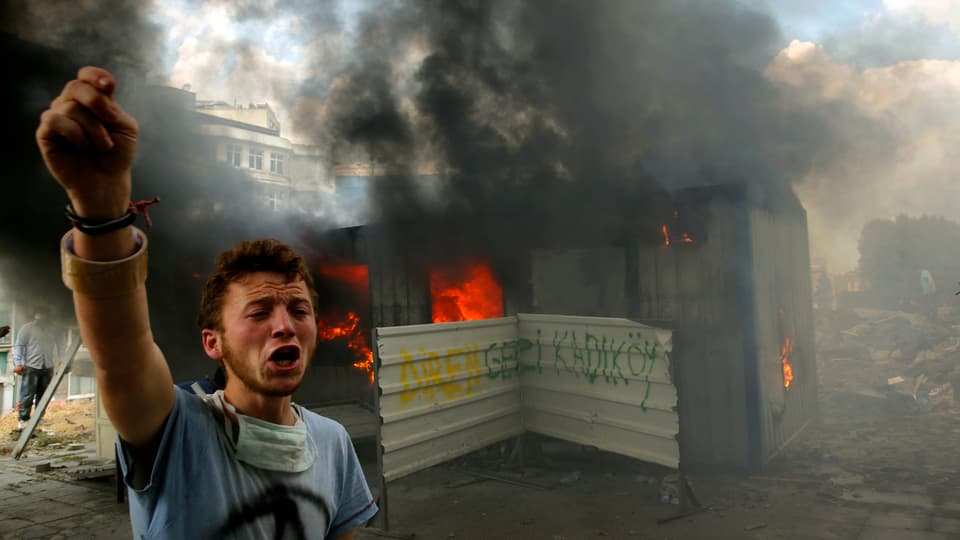 Ein Jugendlicher steht vor einer brennenden Barrikade und schreit, einen Arm erhoben, einen Slogan.