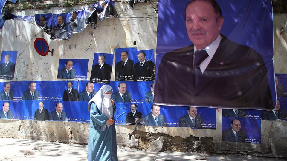 Eine Frau geht vor einer Mauer mit Wahlplakaten von Bouteflika.