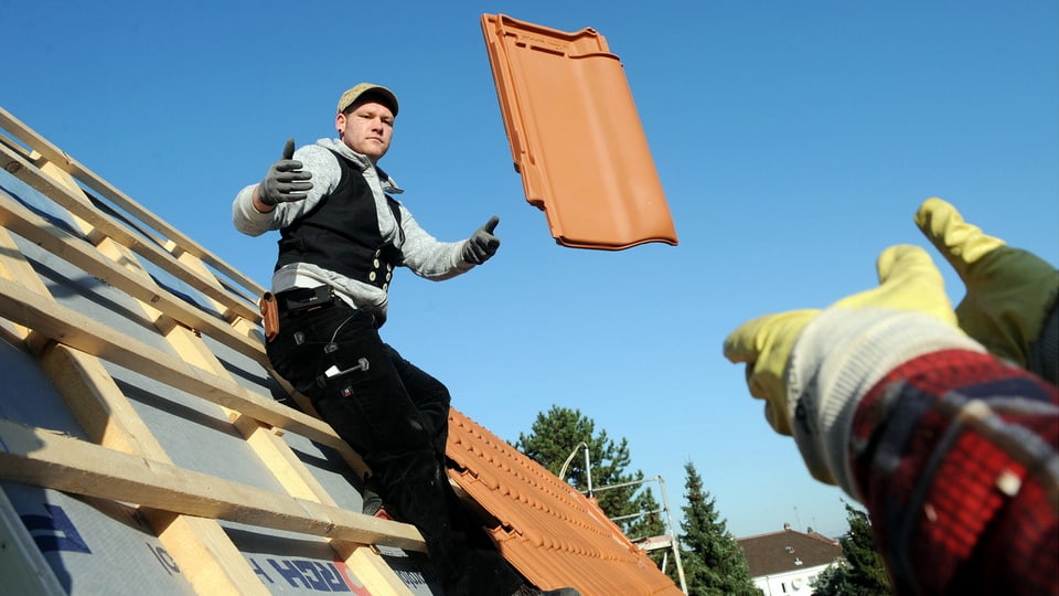 Ein Arbeiter auf einem Dach wirft einem anderen einen Dachziegel zu.