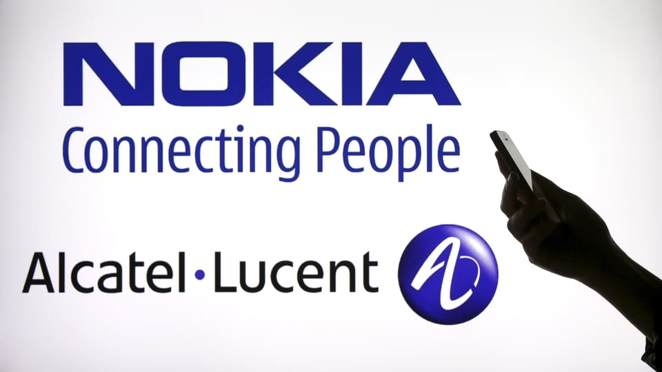 Eine Frau hält ein Handy vor einer Anzeigetafel, auf der steht: Nokia, connecting people. Darunter der Name Alcatel Lucent.