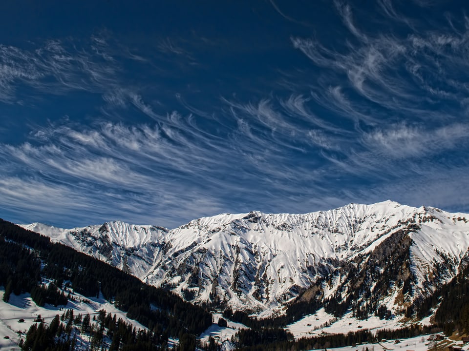 Der Blick ins Tal zeigt grüne Sonnenhänge und schneebedeckte Bergflanken. Am blauen Himmel hat es federartige Schleierwolken.