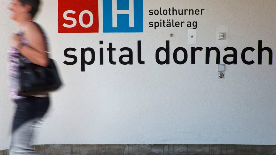 Logo des Spital Dornach an einer Wand, davor eine Passantin