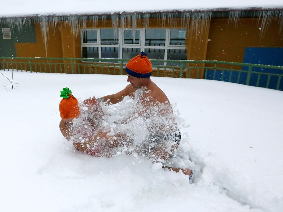 Zwei Männer in Badehose und mit orangen Mützen ringen im Freien bei Neuschnee.