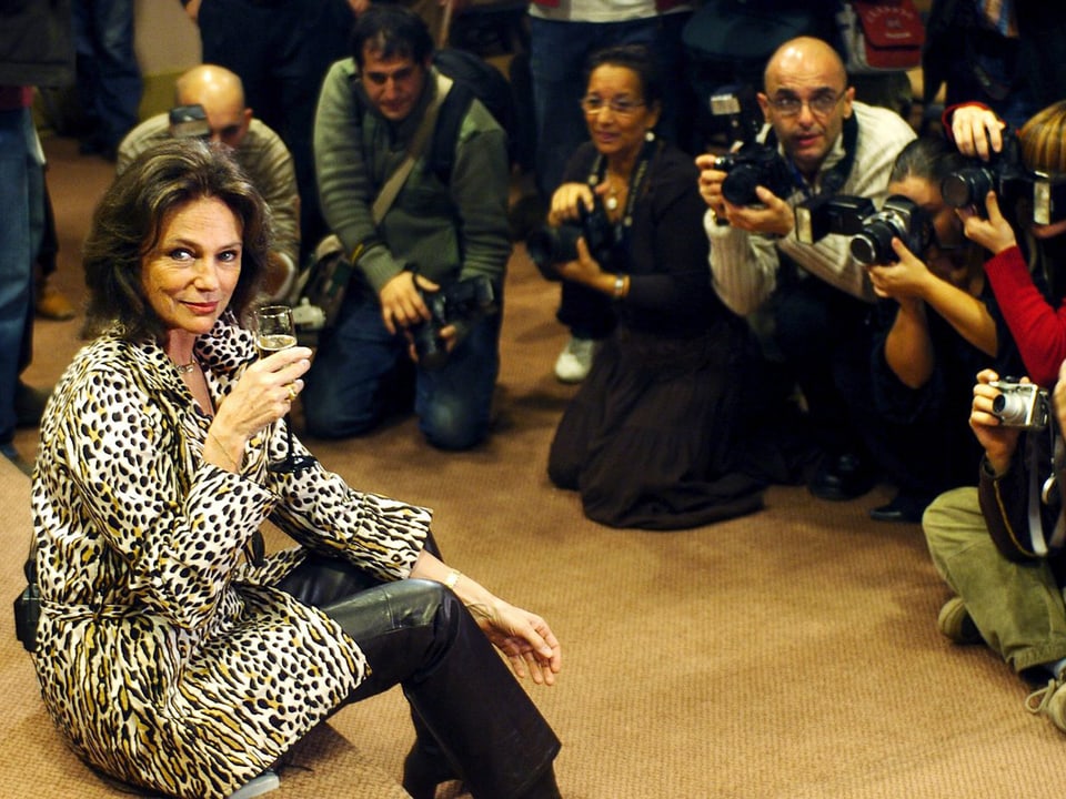 Eine Frau im Leopardenmantel und Sektglas in die Hand sitzt auf einer Stufe umringt von Fotografen.