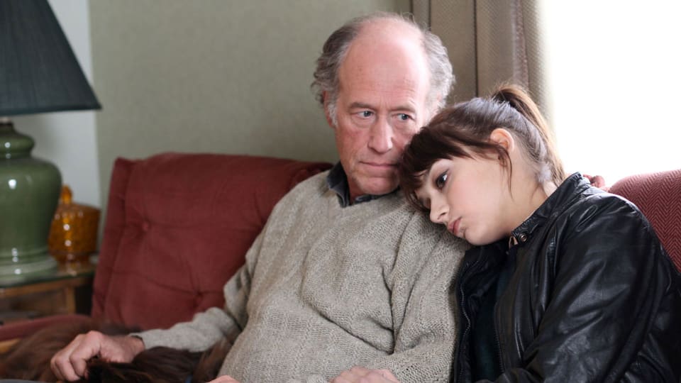 Ein älterer Mann sitzt auf dem Sofa, neben ihm eine junge Frau, die ihren Kopf an seine Schulter lehnt.