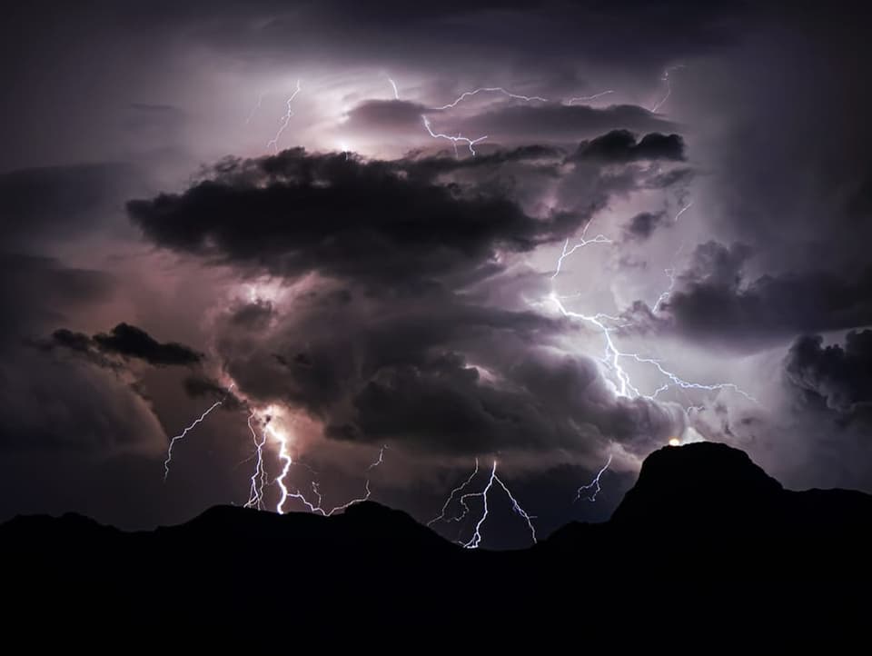 A dark night with storm clouds over the borders of the mountains, lit by only a few lightning bolts.