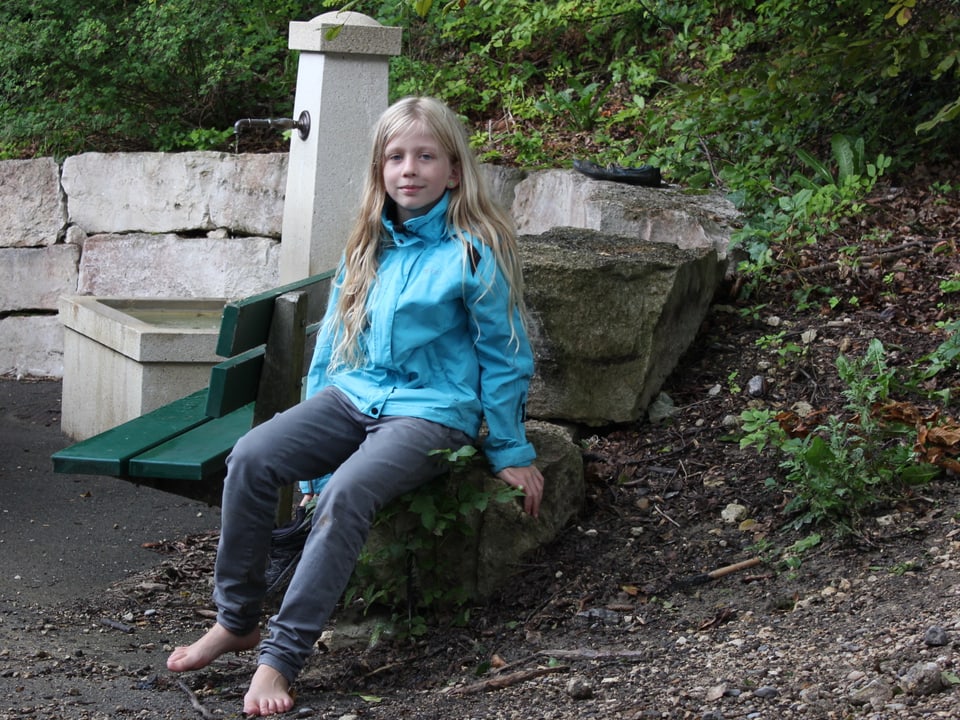 Mädchen sitzt barfuss auf einem Stein bei einem Brunnen.
