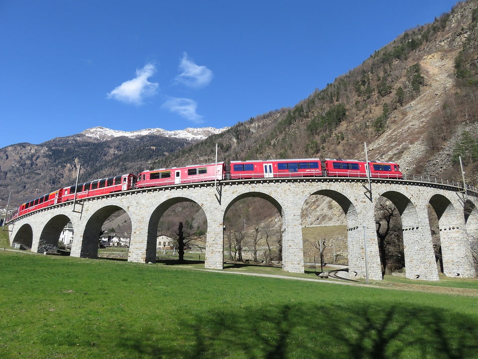 Ein roter Zug der Rhätischen Bahn befährt ein aufsteigendes Kreisviadukt. Im Hintergrund sind Berge und blauer Himmel.