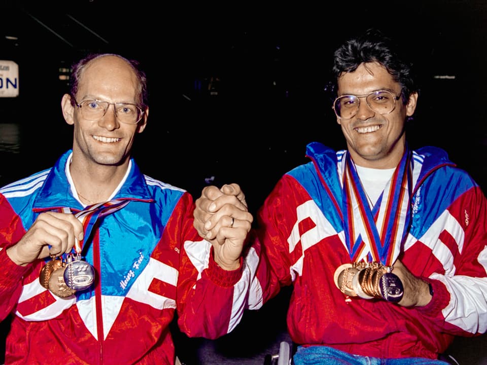 Zwei strahlende Männer mit vielen Medallien um den Hals in altertümlich wirkenden Trainingsanzügen. 
