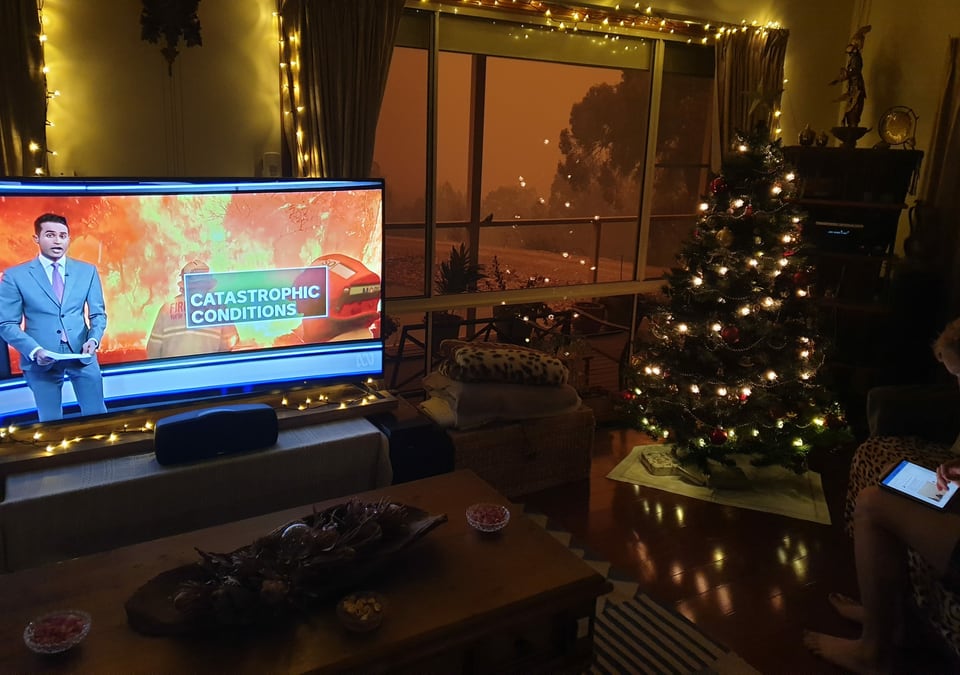 Blick auf einen Fernseher, der über die Brände in Australien berichtet, durch das Fenster ist der Himmel zu erkennen, orange von den Feuern.