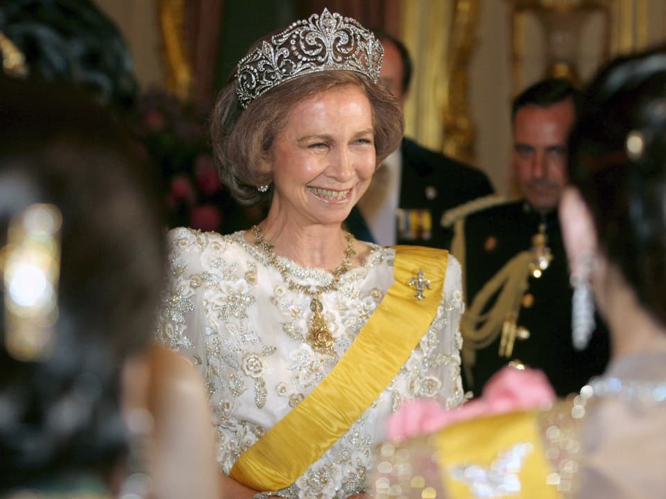 Königin mit Krone und weissem Kleid steht in der Menschenmenge und lächelt.