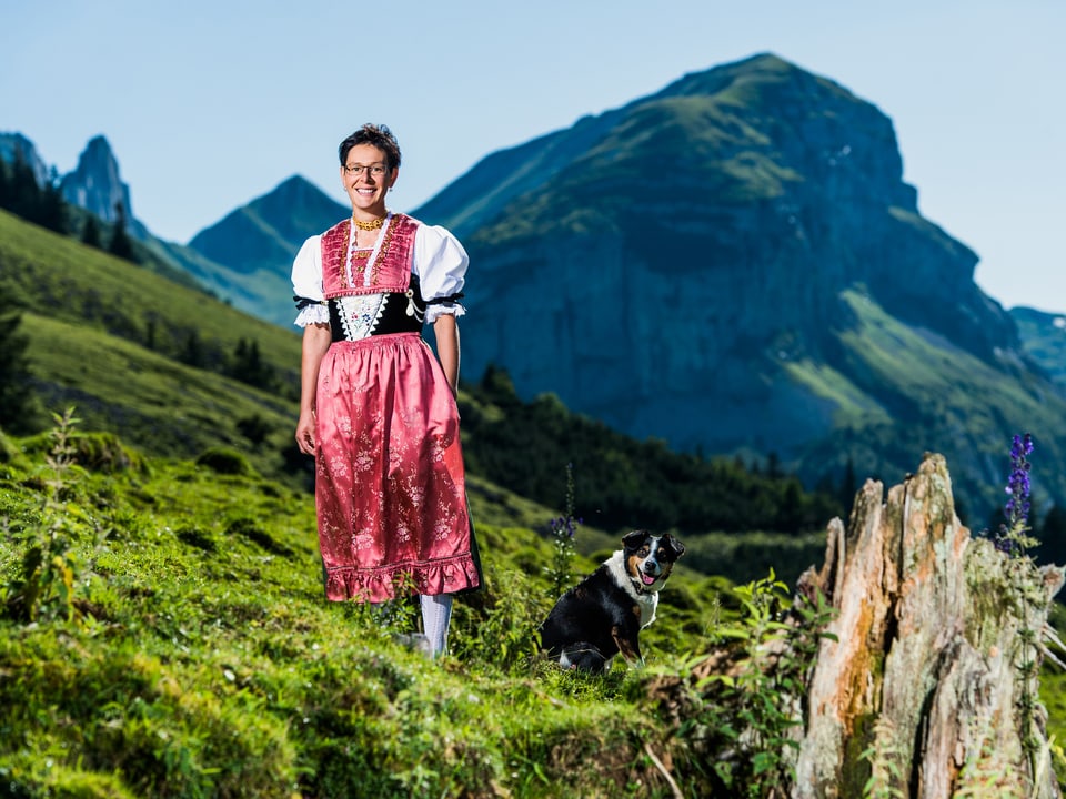 Theresia Hollenstein steht mit ihrer Tracht auf einer Bergwiese mit dem Hofhund daneben.