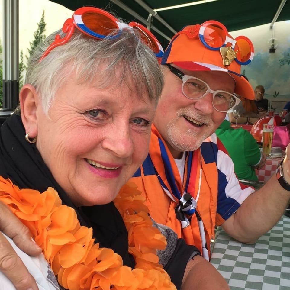 Älteres Ehepaar lächelt vergnügt in die Kamera, die beiden tragen orange Fanutensilien aus Holland. 