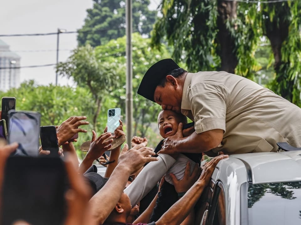 Prabowo küsst ein schreiendes Baby aus einem Auto heraus. Dicht gedrängt um das Auto herum stehen viele Menschen.