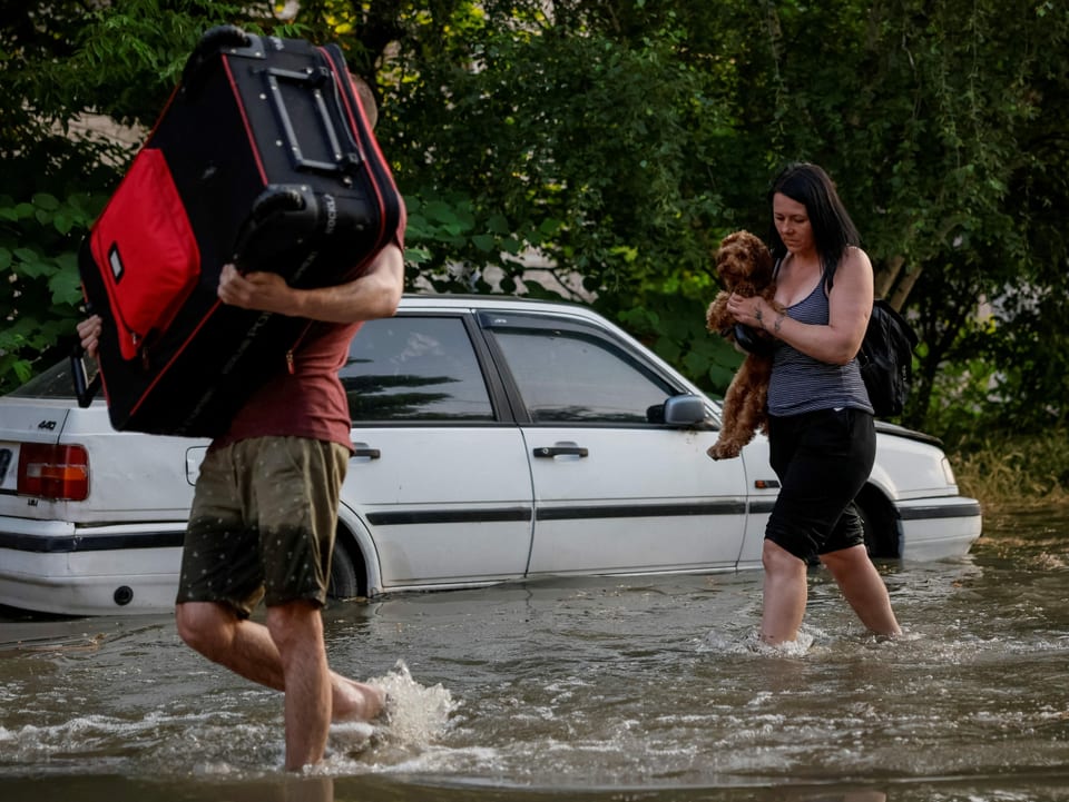 Ein Mann trägt eine Koffer und eine Frau trägt einen Hund. Sie laufen auf einer überschwemmten Strasse.