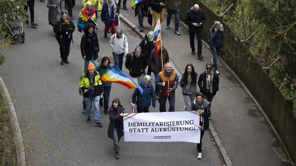 Menschen tragen Plakat und Regenbogenfahnen. Auf Plakat steht: Demiliatirisierung statt Aufrüstung.