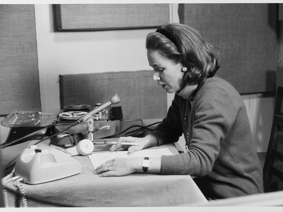 Eine Frau sitzt an einem Tisch, vor sich ein Mikrofon und ein Telefon - der Hörer liegt daneben.