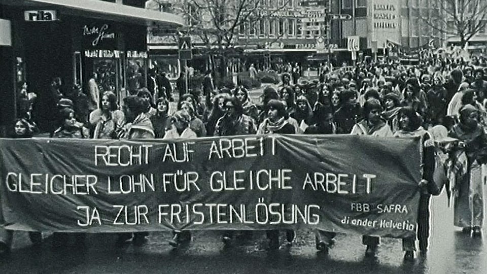 Demonstration. Strasse voll mit Menschen. Vorne tragen sie ein Plakat auf dem steht: Recht auf Arbeit! Gleicher Lohn für gleiche Arbeit. Ja zur Fristenlösung. Das Foto ist schwarzweiss.