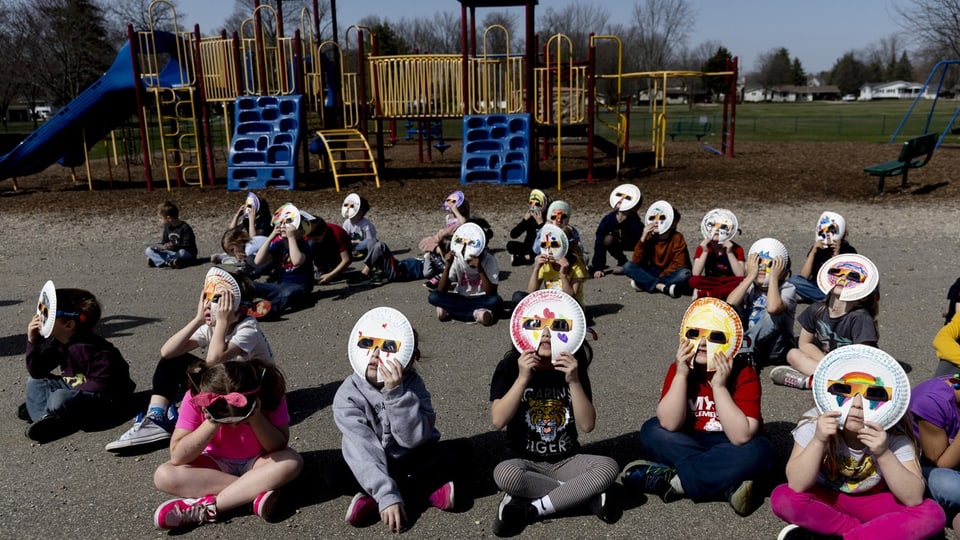 Kinder auf dem Schulhof sitzen auf dem Boden. Sie halten gebastelte Masken mit Sonnenfinsternisbrillen vors Gesicht. 