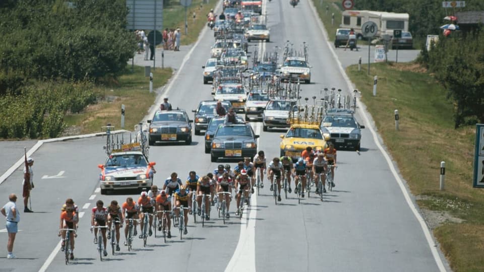 Eine Gruppe von professionellen Rennradfahrern vor einer Autokolonne.