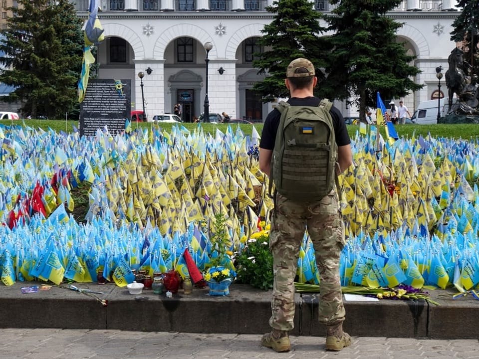 Ein ukrainischer Soldat steht vor einem Denkmal das mit Flaggen gefallener Soldaten geschmückt ist.