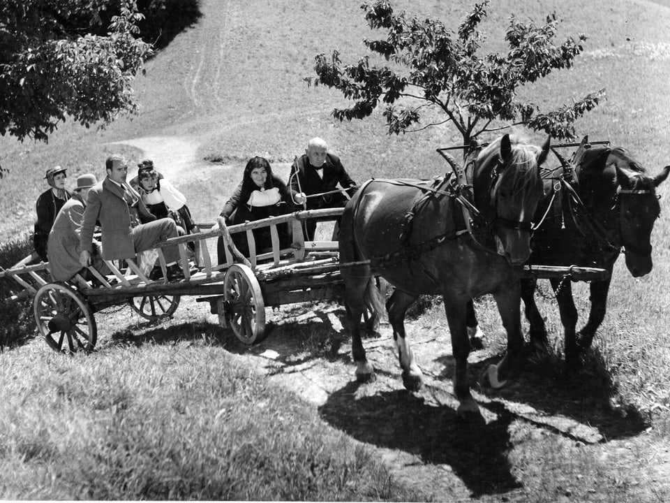 Zwei Pferde ziehen eine grossen Leiterwagen mit drei Frauen und drei Männern einen steilen Weg hinauf.