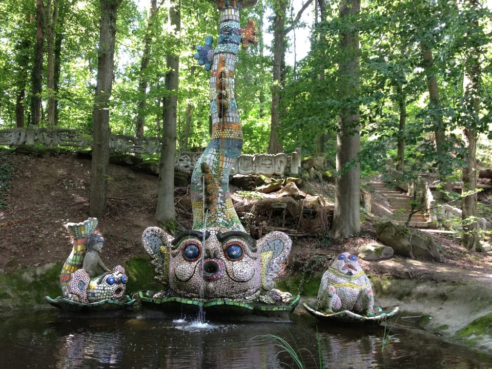 Ein wasserspeiendes Wesen an einem Teich im Wald, flankiert von zwei kleineren Skulpturen