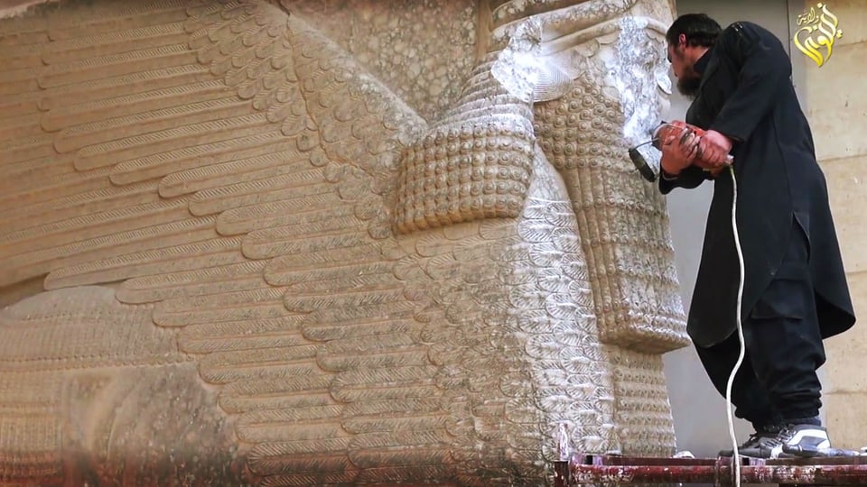 Ein schwarz bekleideter Mann zerstört mit einem Presslufthammer eine grosse Figur aus Stein.