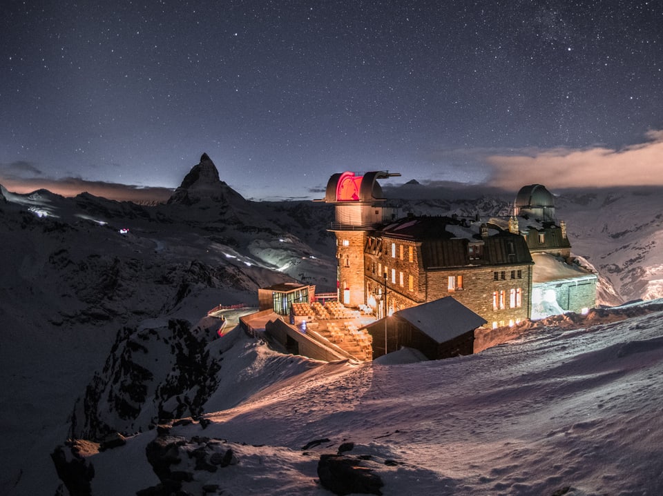 Foto bei Nacht mit eher hellem Sternenhimmel. Alpen mit Bergen und Gipfeln im Schnee, im Vordergrund ein beleuchtetes Gebäude mit Kuppeln, die Sternwarte. 