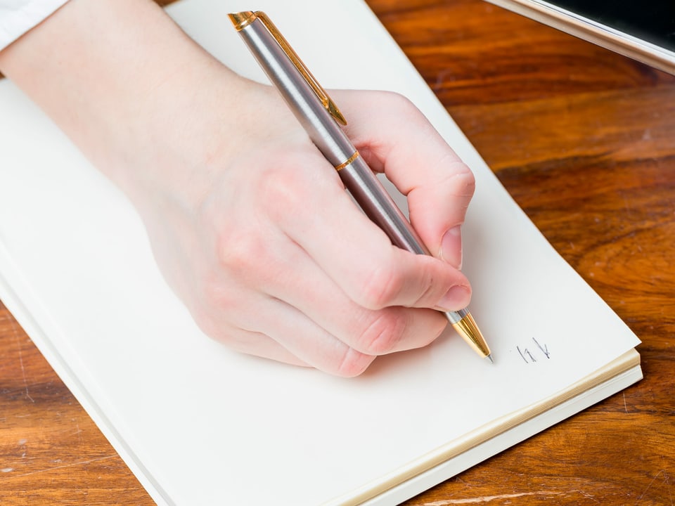 Eine Frauenhand mit Kugelschreiber beginnt einen Brief auf einem Schreibblock.