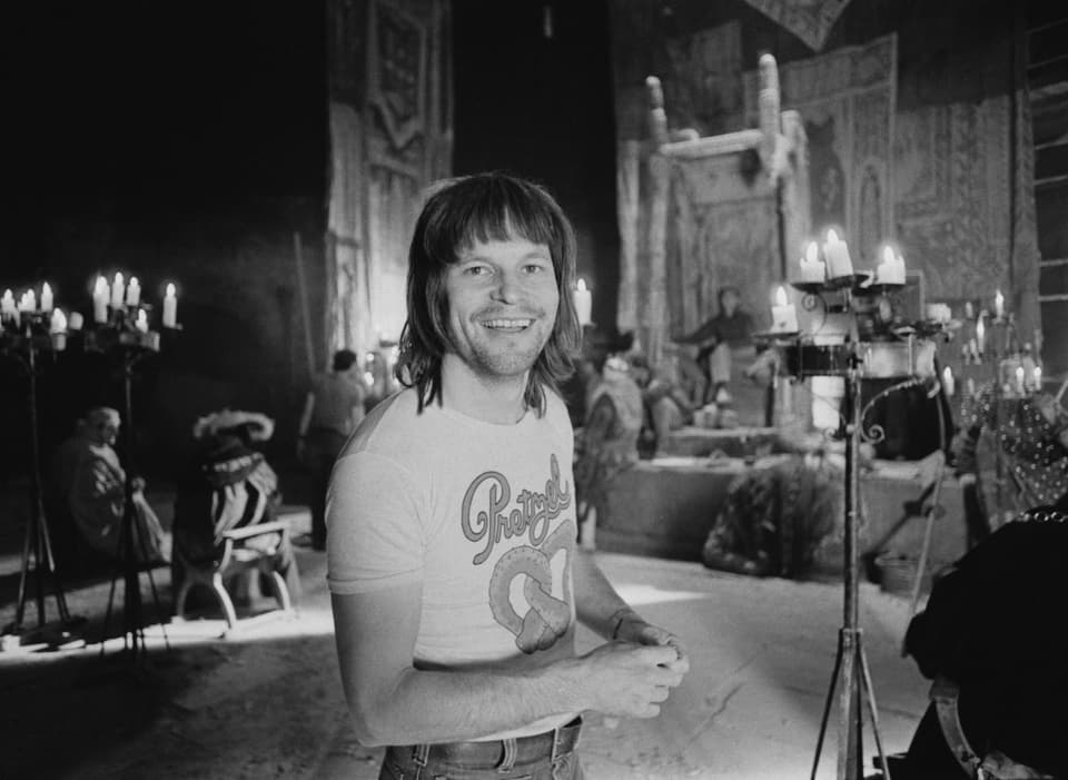 Ein Mann mit halblangen Haaren und einem Print-Shirt in einer Kirche lächelt in die Kamera.