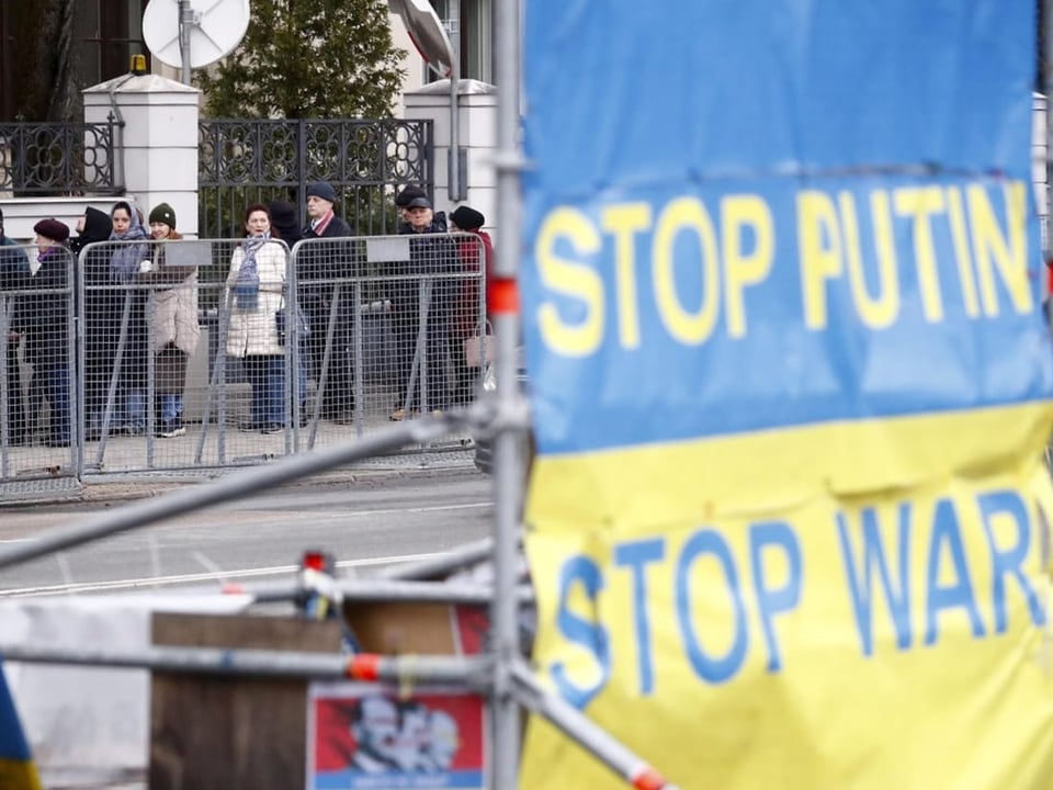 Im Hintergrund: Menschenschlange. Im Vordergrund ukrainische Flagge mit der Aufschrift «Stop Putin – Stop War»