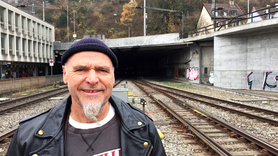 EIn Mann mit Ziegenbart, Lederjacke und Wollmütze steht lächelnd auf einem Bahngleis.