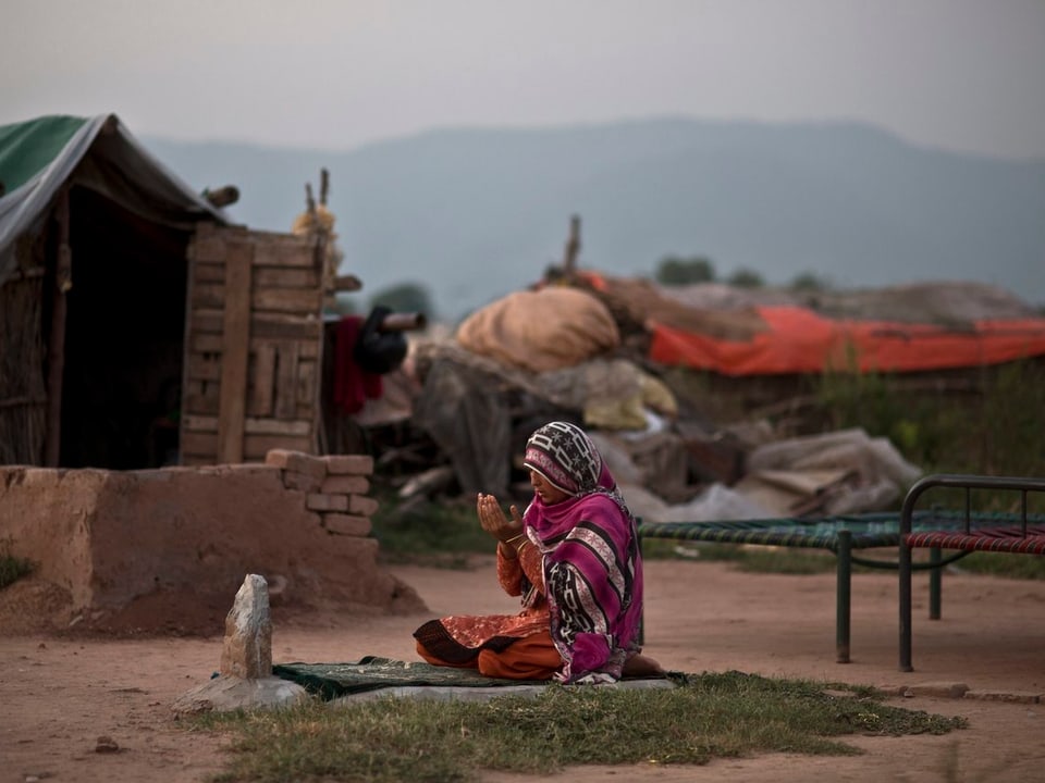 Eine junge Frau kniet am Boden und betet, sie trägt einen pinken Sari. Im Hintergrund eine ärmliche Umgebung, Lehmhütten und kaputte Liegen. 