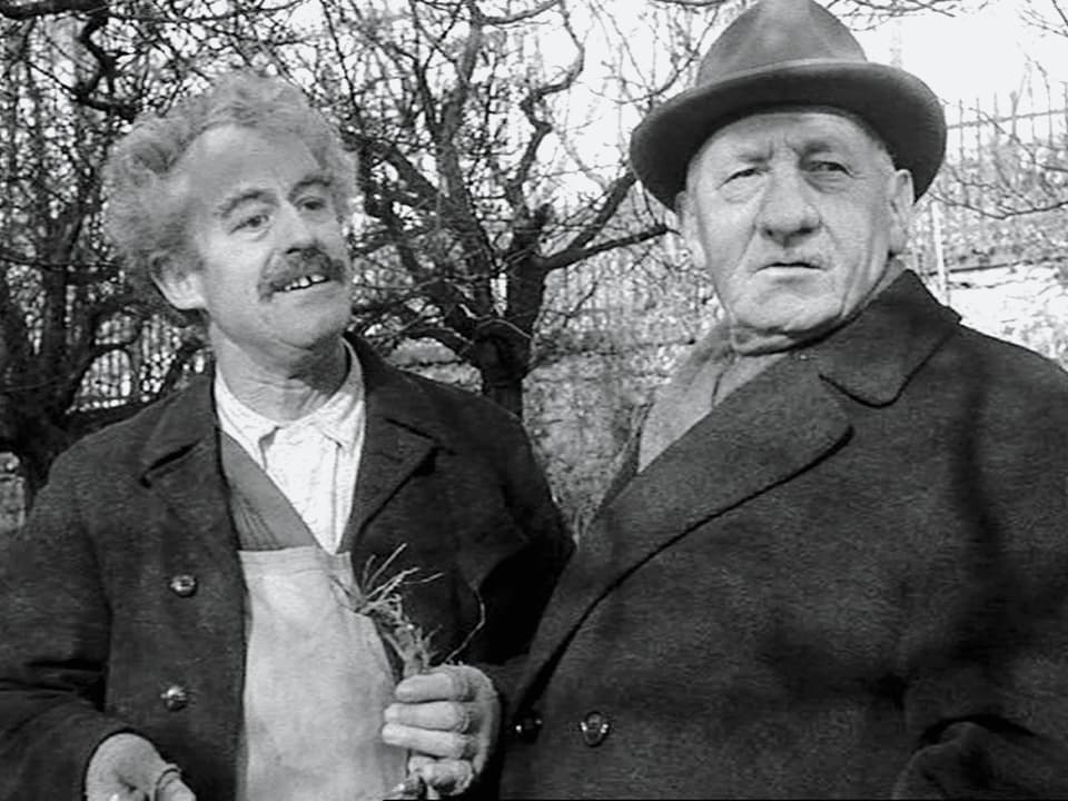 Ein Mann mit Schürze hält eine Pflanze in der Hand und steht neben einem älteren Mann mit Hut.