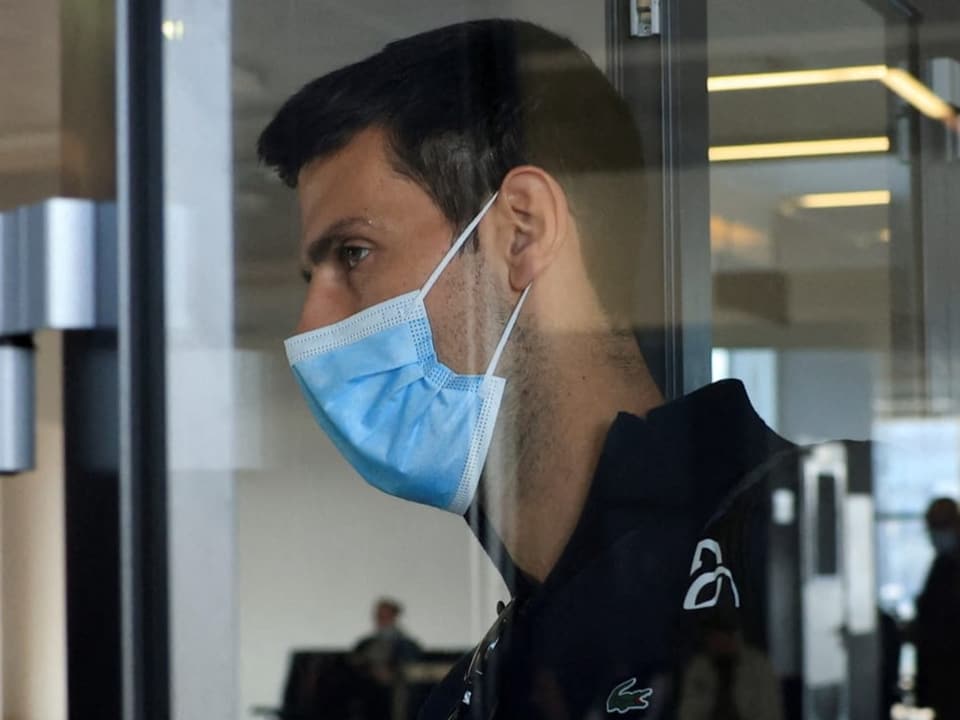 Djokovic geht mit Maske durch eine Glastür am Flughafen.