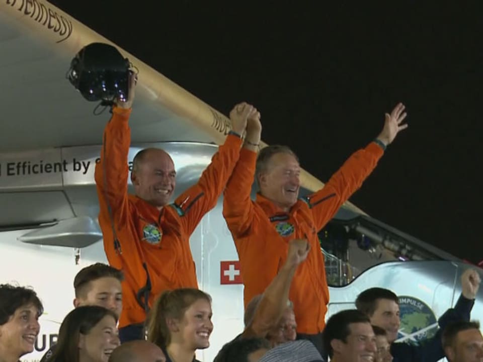 Zwei jubelnde Piloten stehen vor einem Solarflugzeug. Im Fordergrund eine Gruppe von lachenden Leuten.