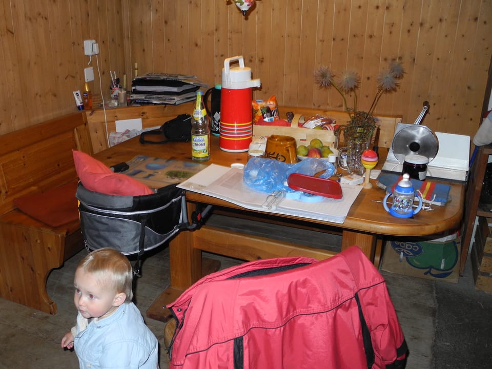 Holztisch mit hölzerner Eckbank. Auf dem Tisch stehen Diverse Gegenstände wie Thermoskanne, Spielzeug, Vase oder eine Früchteschale.