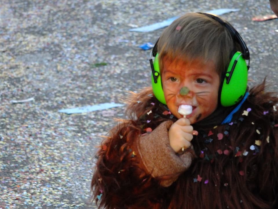 Kind in Bärenfell mit Gehörschutz und Schleckstengel.